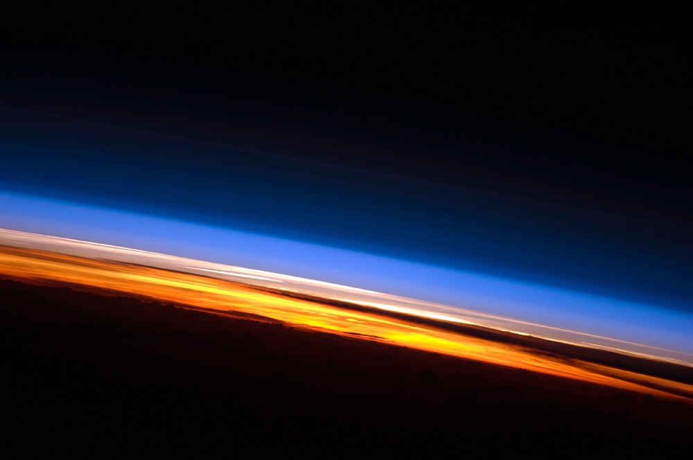 Orange, white and blue atmospheric bands above Earth’s dark curved edge at sunset